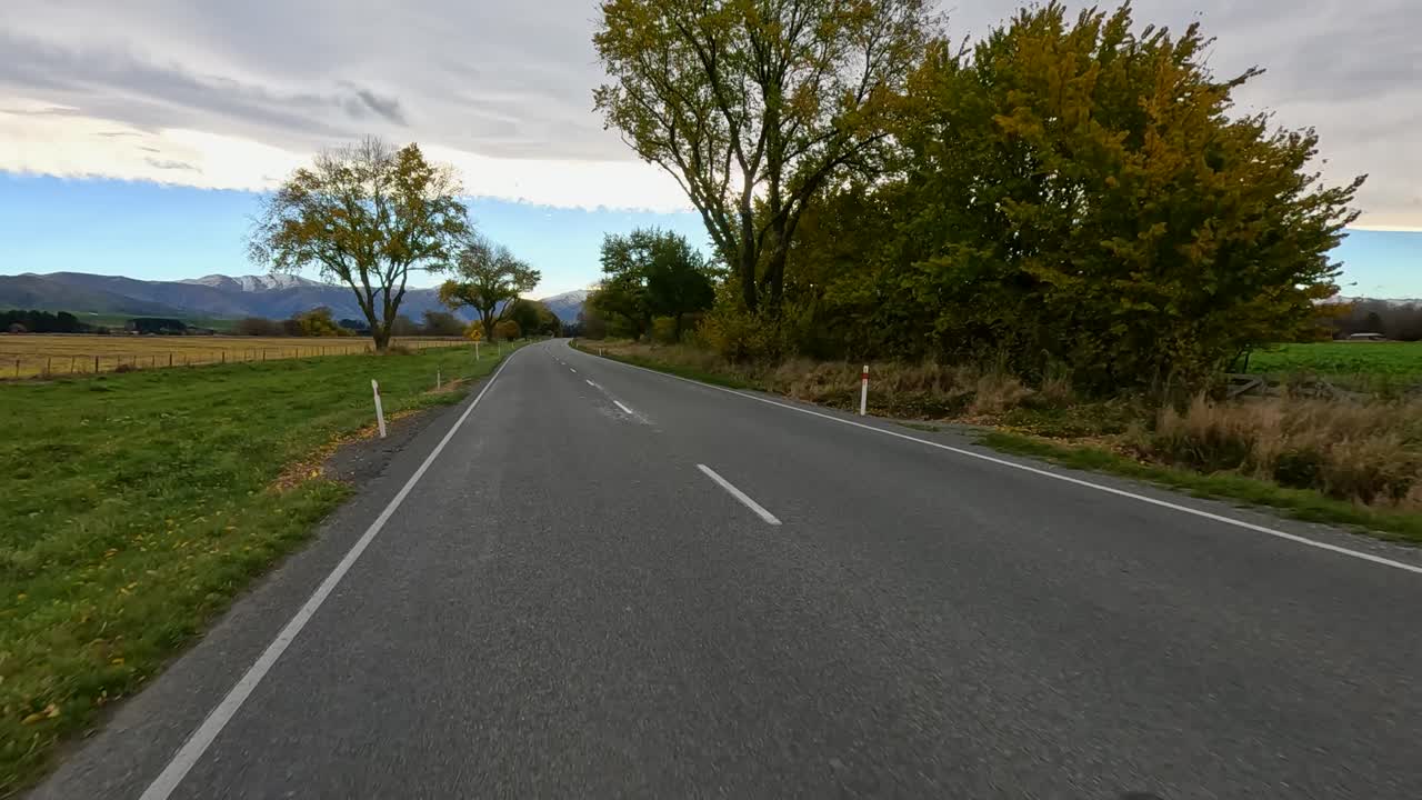 A smooth forward drive along a rural road lined with autumn trees, under overcast skies, with distant mountains visible. Static, wide-angle, natural lighting