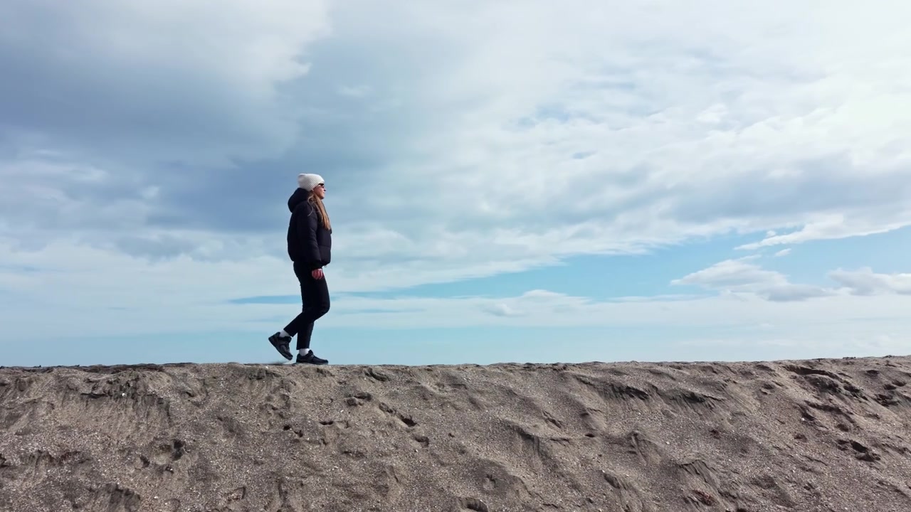 Woman walks on a sandy path by the ocean under a cloudy sky in a coastal location during the day