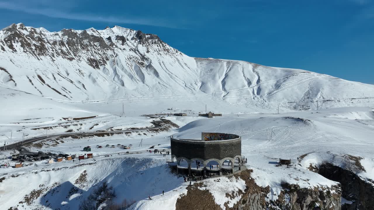 A grand circular monument sits atop a snow-covered plateau, surrounded by vast, untouched terrain