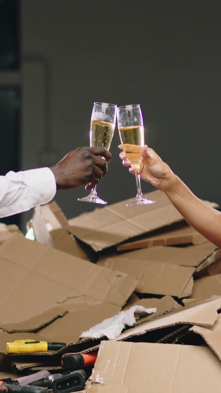 Vertical video: Toasting flutes after moving at home, couple with white shirt over boxes and tools