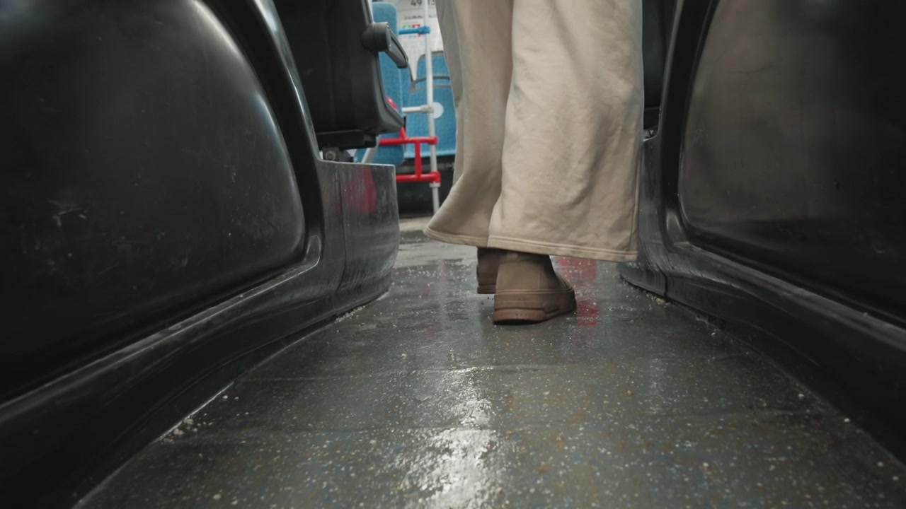 Prospettiva posteriore dal basso di una persona che cammina lungo uno stretto corridoio di transito verso i sedili distanti, con sollevamento visibile del tallone, trasferimento del peso in avanti, scarpe usurate, pavimento strutturato, luce soffusa e forte