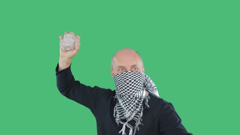 anarchist in checkered bandana swings stone om green background on demonstration. bald vandal with stone on civil revolution. social riots and uprisings concept.