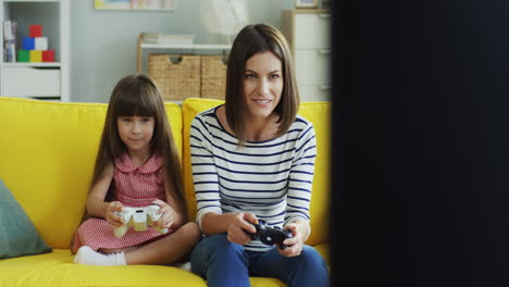 Alegre-Joven-Madre-E-Hija-Jugando-Videojuegos-Con-Joystick-Mientras-Se-Sientan-En-El-Sofá-En-La-Sala-De-Estar