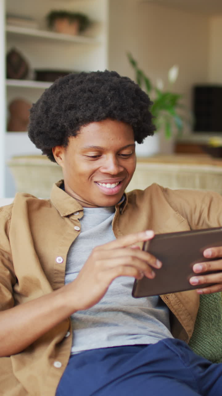 Vertical video of happy african american man using tablet and laughing