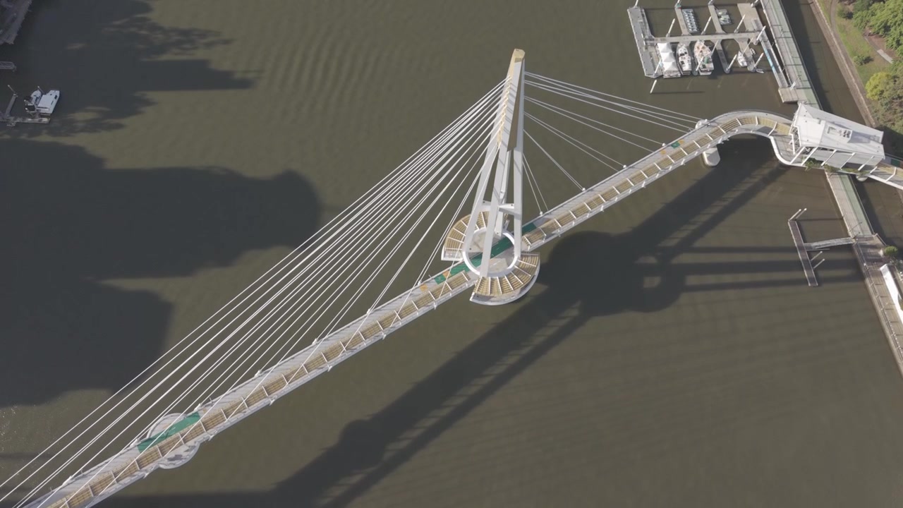 A top down aerial view looks along the Kangaroo Point Bridge pylon and cables, a maintenance boat visible on the river below.