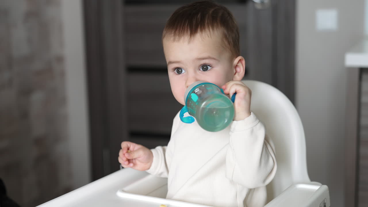 Little toddler sitting at the feeding table reaches hand for the bottle. Mom's hand gives a bottle, kid drinks water and puts it on the table. Close up.