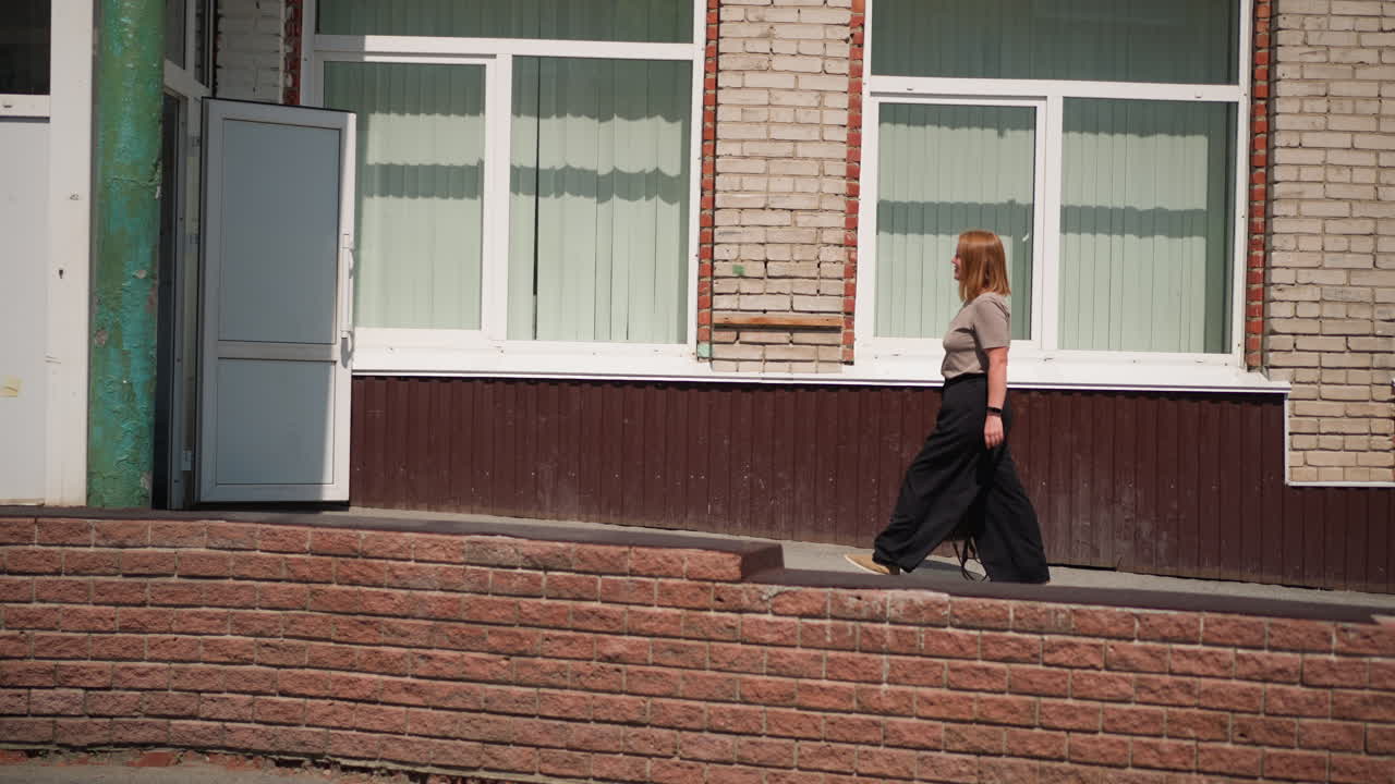Side view of teacher walking into building carrying handbag under bright sunlight, brick wall and large windows reflecting calm academic environment