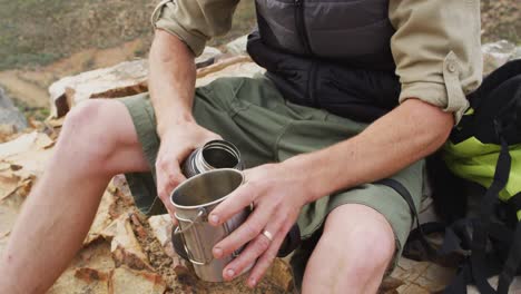 mid section of caucasian male survivalist sitting on mountain in wilderness, pouring water to drink