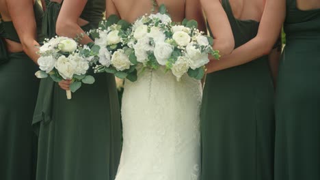 camera pans up from dresses to show bridesmaids and bride holding bouquets in a garden