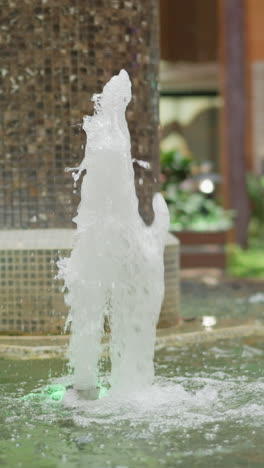 fuente de agua interior con arroyos de agua iluminados por luces de colores en un centro comercial moderno, rodeado de pilares de azulejos, escaleras mecánicas y vegetación