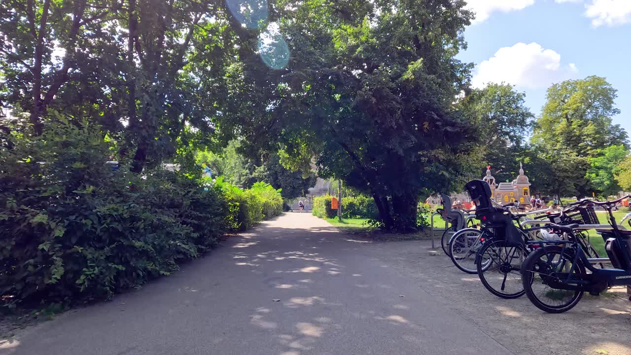 A steady daytime walk along a tree-lined park path in Berlin, passing bicycles, greenery, and people under bright natural sunlight with gentle camera movement