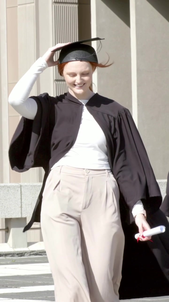 Video verticale: Una neolaureata con diploma si stabilizza mentre il vento le solleva la toga e il tocco, camminando in piazza.