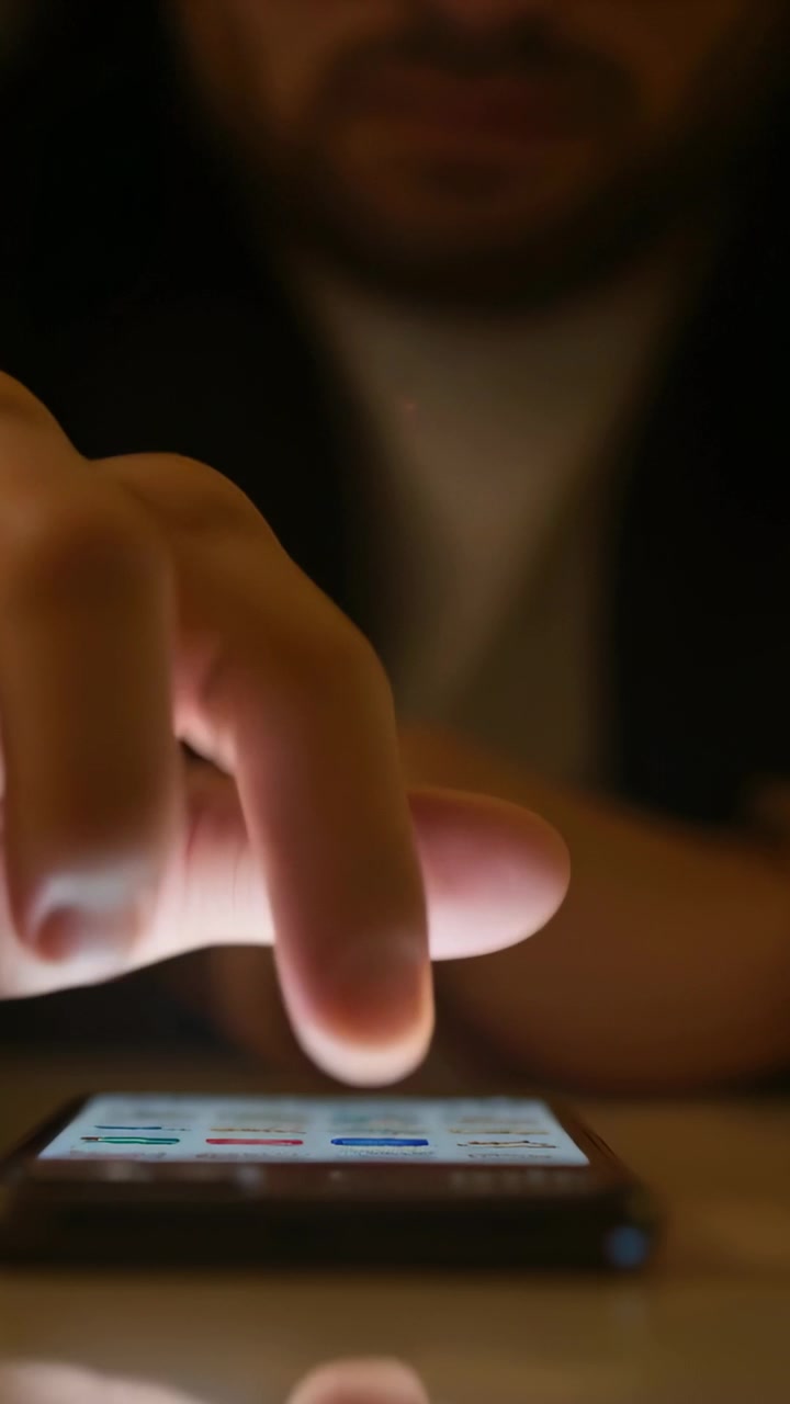Dito che illumina il telefono toccandolo, uomo con giacca scura che scorre sul tavolo in una stanza in penombra, navigando tra le app.