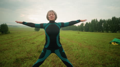 woman in green and black suit on yoga mat practicing triangle pose with arms and legs extended, standing in an open grassy field, surrounded by greenery and trees under a cloudy sky