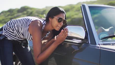 african american woman applying lipstick while looking in the mirror of convertible car on road