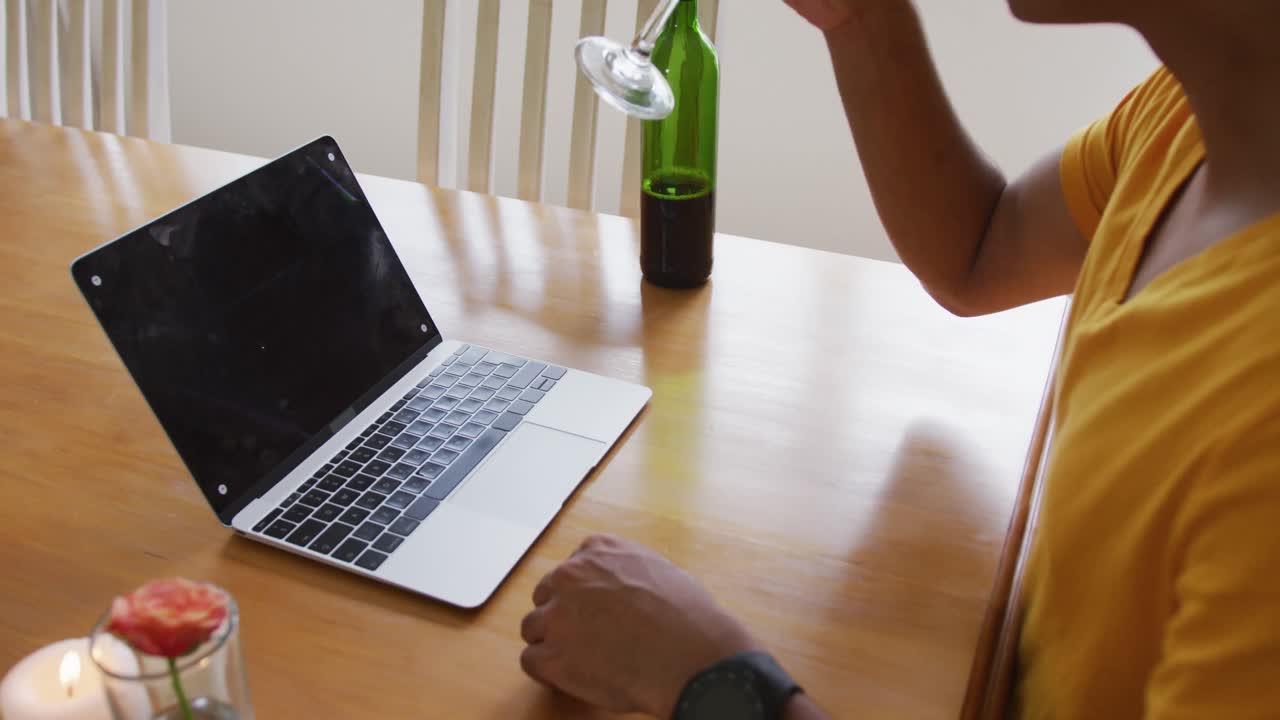 Midsection of mixed race man sitting at table making video call with laptop drinking wine and waving