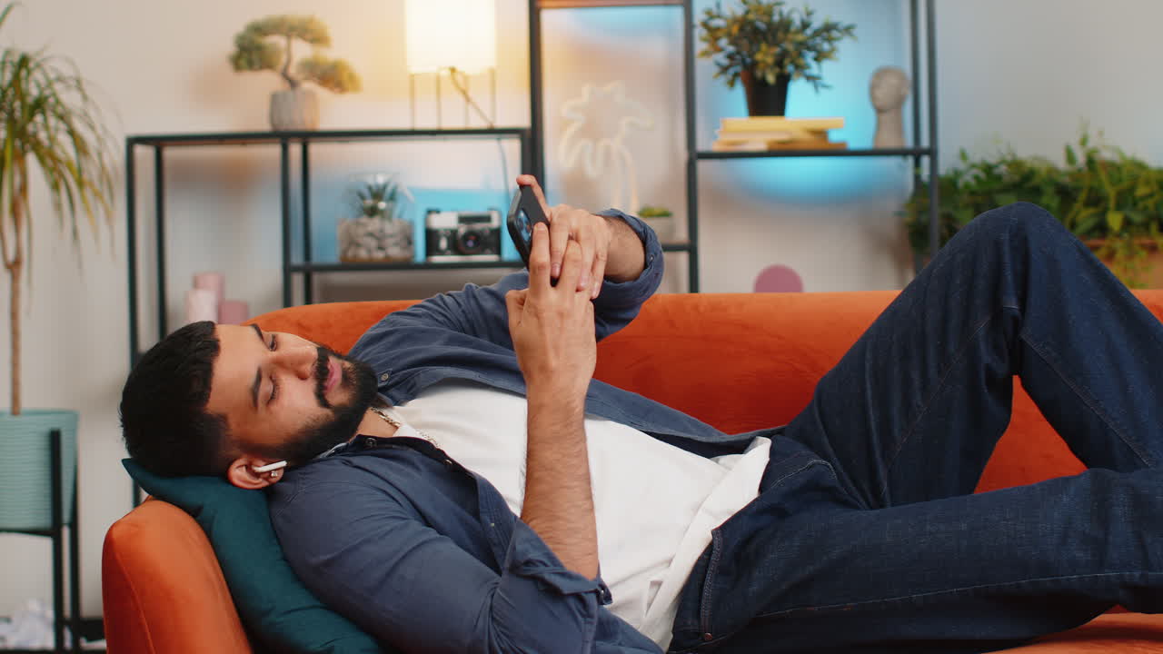 Indian man taking wireless earphones out of box listening music enjoying leisure relaxing at home