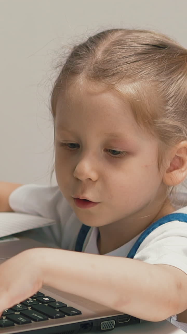 closeup slow motion little girl student sits at table with notebook laptop, blonde with pigtails child bored reasoning, presses buttons on keyboard
