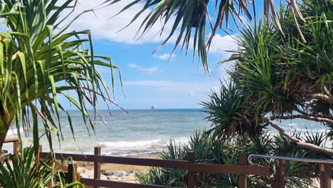 A-gorgeous-blue-beach-that-harbors-a-boat-in-the-background-sits,-hidden-behind-beach-trees-and-a-wooden-railing-on-a-perfect-sunny-day-where-fluffy-white-clouds-drift-overhead