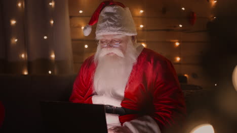 Der-Weihnachtsmann-Arbeitet-Am-Heiligabend-Zu-Hause-Mit-Einem-Laptop.-Weihnachtsbeleuchtung-Und-Dekoration