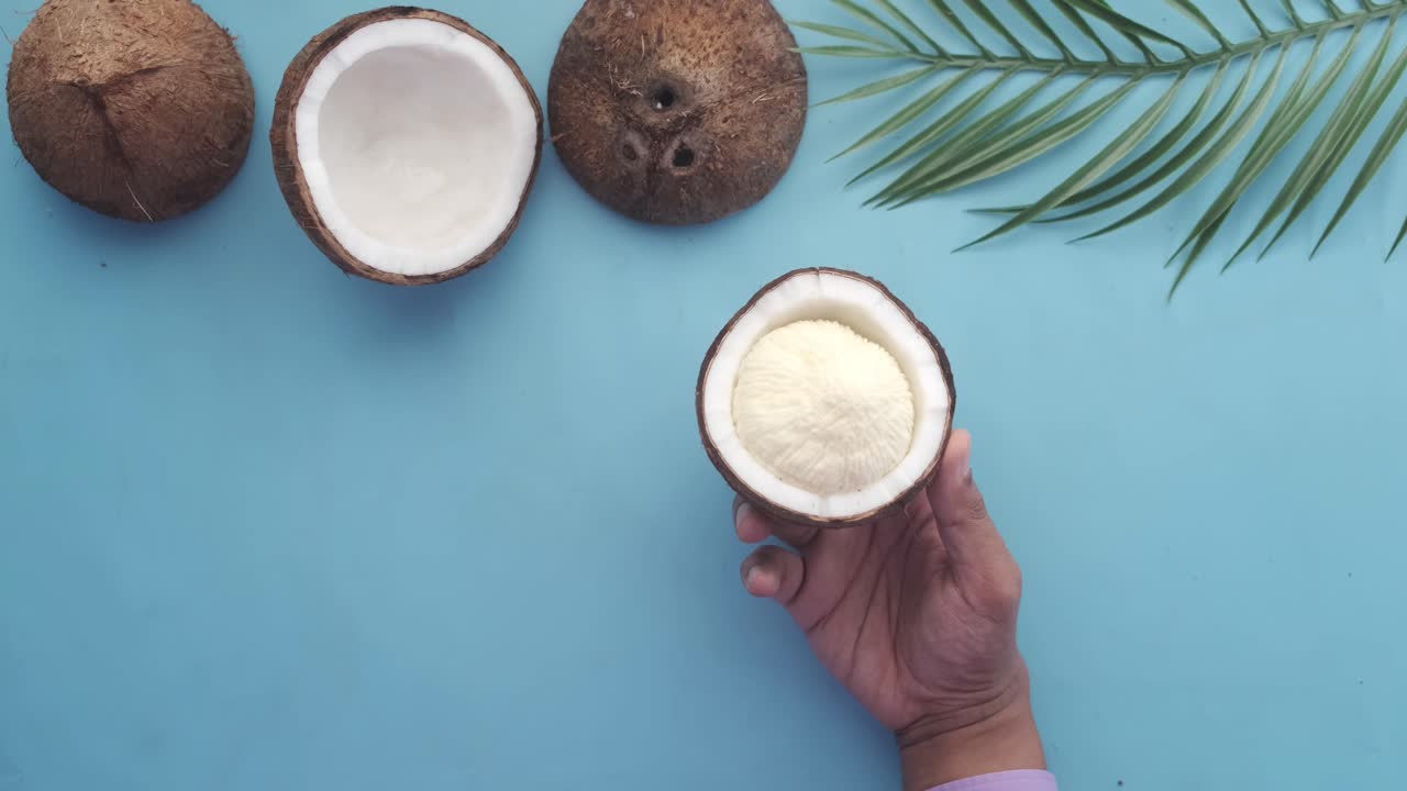 mano sosteniendo un coco cortado en un fondo azul claro con otros cocos y hojas de palma
