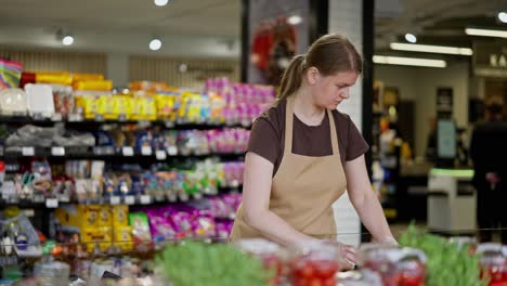 Confident-girl-in-a-brown-T-shirt-and-apron-lays-out-vegetables-and-fruits-on-the-counter-in-a-supermarket-during-her-working-day