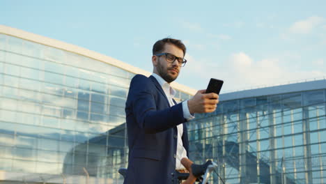 buen empresario tomando una foto selfie mientras está de pie con una bicicleta fuera de un edificio moderno