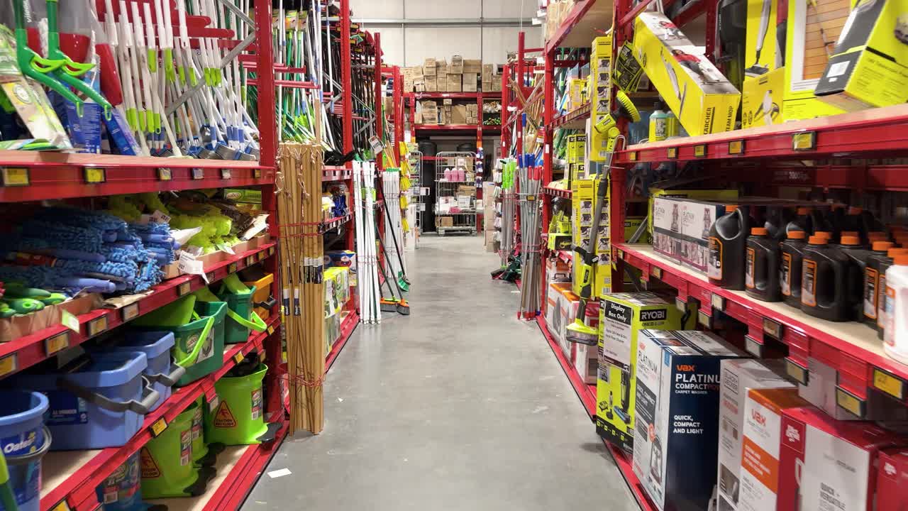 Camera moves through hardware store aisle stocked with cleaning tools, bright lighting, organized shelves