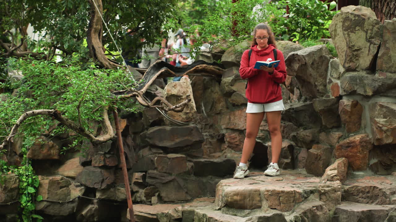 Giovane ragazza bianca con giacca rossa controlla la fotocamera sui gradini rocciosi di un giardino, in piedi su una sporgenza di pietra accanto a un muro muschioso e alberi frondosi. Indossa pantaloncini e scarpe da ginnastica, mentre rivede le foto e filma un vlog di viaggio.