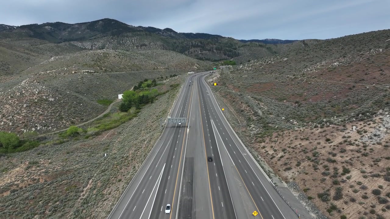 Autostrada a più corsie del Nevada che si estende attraverso un paesaggio desertico arido con colline rocciose, vegetazione rada e arbustiva e una catena montuosa aspra sotto un cielo azzurro parzialmente nuvoloso. Nevada, USA. Ampia ripresa aerea in ascesa.