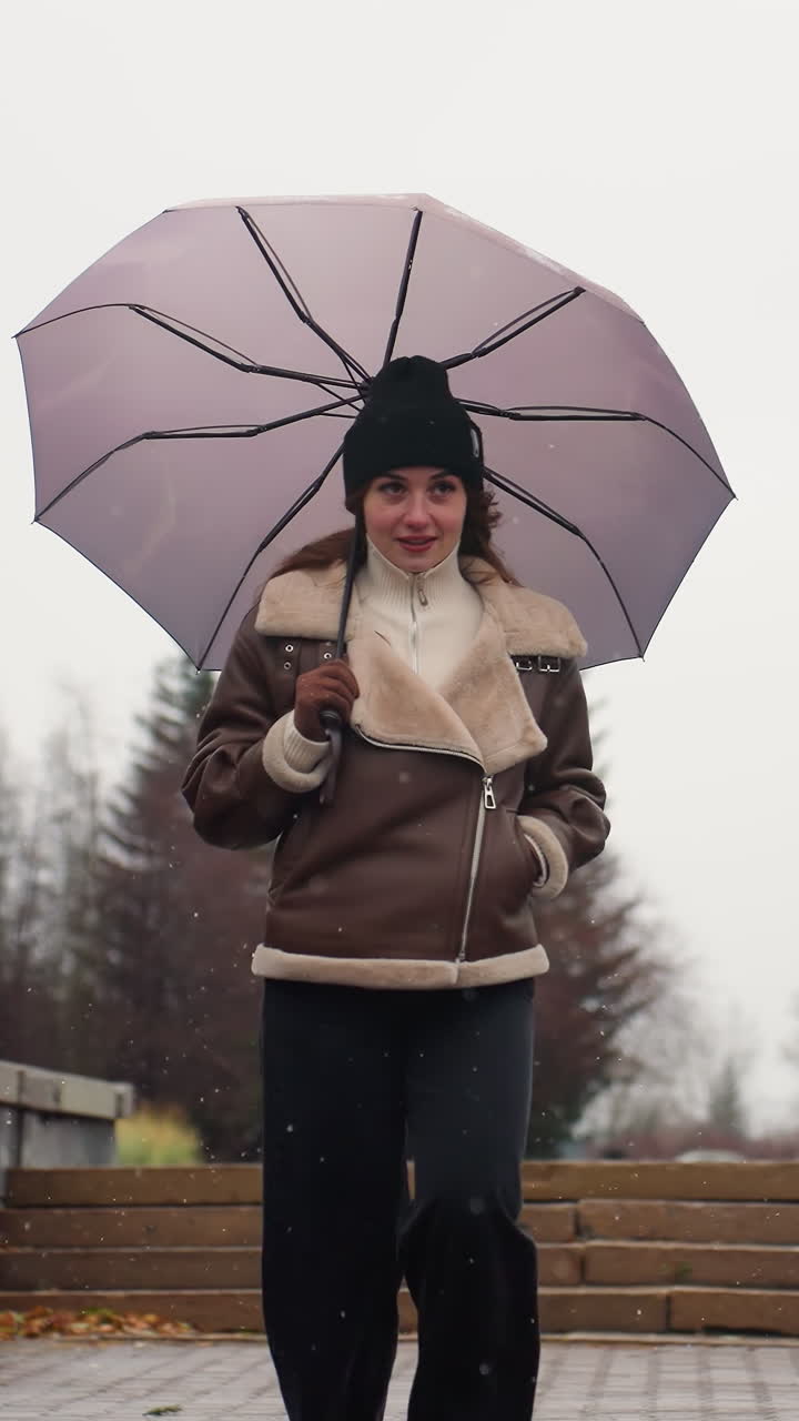 Young woman running in light snowfall holding umbrella wearing knit cap brown shearling jacket black trousers one hand pocket urban background autumn leaves cityscape overcast trees buildings pavement