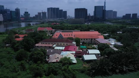 Thủ-Thiêm-Parish-Church-and-the-Lovers-of-the-Holy-Cross-Convent-are-the-oldest-French-Colonial-buildings-in-Ho-Chi-Minh-City,-Vietnam-Aerial-view-orbitting-settlement-revealing-modern-city-skyline