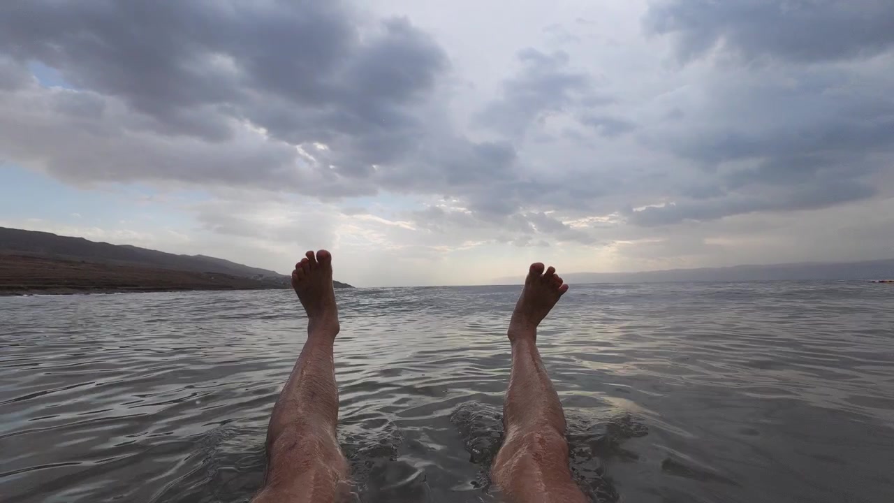 POV di un uomo che galleggia con gambe e piedi sollevati nel Mar Morto ipersalino della Giordania