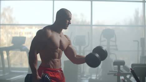 a man with a naked torso and a pumped strong body and relief muscles of the press raises dumbbells against the background of the window training his hands. strength of will and stubborn character.