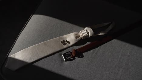 groom's white tie and brown leather belt laid out on a bed in sunlight