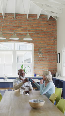 vertical shot of a senior biracial couple drinking coffee at a dinner table, laughing and smiling
