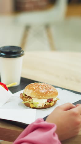 vista trasera de una mujer con vestido rosado en una mesa, comiendo de una bandeja negra que contiene una hamburguesa, papas fritas y café, ella recoge una patata frita de las papas frites y disfruta de su comida con una postura relajada