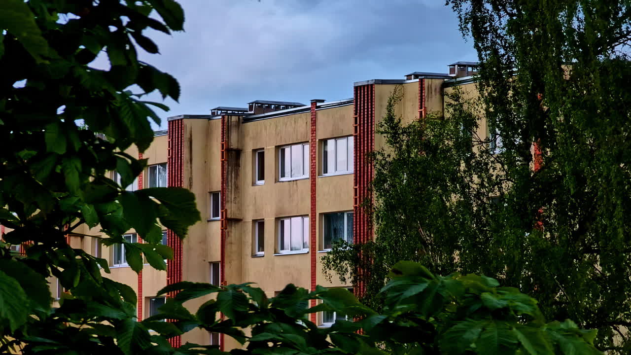 Old Soviet-era apartment building with overgrown trees and cloudy sky in summer