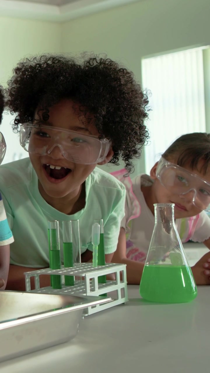 Vertical video: Leaning curly kid wearing goggles in mint top observing green tubes reacting at lab