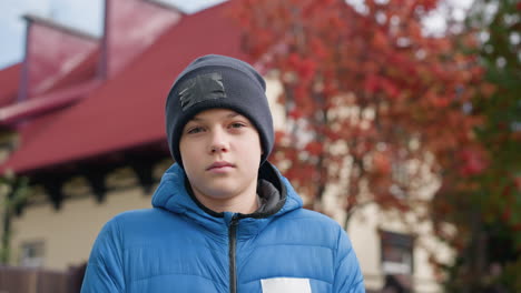 young boy wearing black beanie and blue jacket standing outdoors, looking thoughtfully in front of beautiful autumn trees and house with colorful red foliage in background