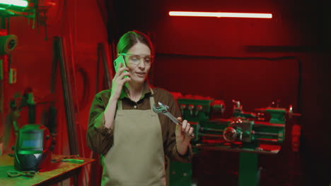 alegre soldadora en delantal y gafas hablando por teléfono en el trabajo en el taller