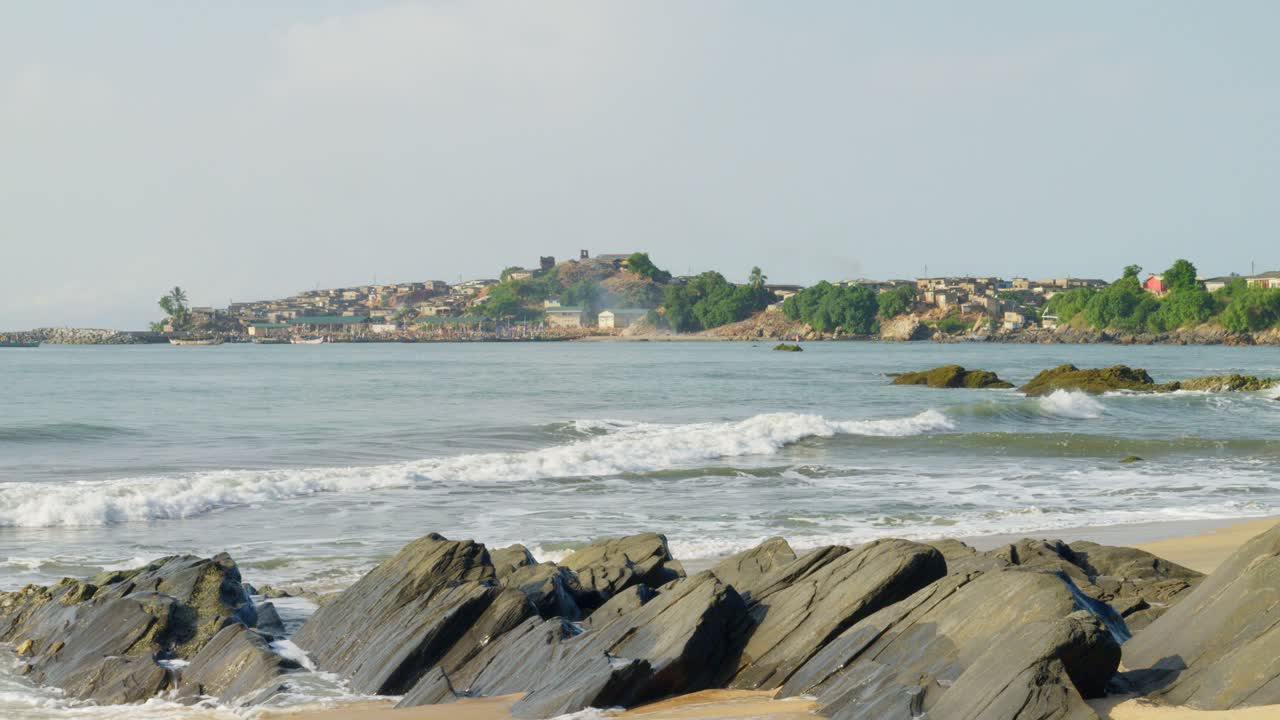 Ondas batendo em praia de areia com rochas negras, cidade costeira além da baía.