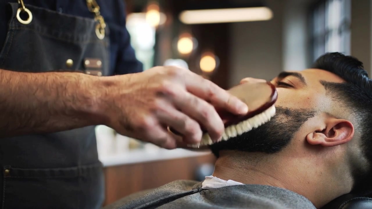 Ein Barbier in Schürze pflegt die Kieferlinie eines zurückgelehnten Kunden mit einer Bartbürste im Geschäft, der Kunde trägt einen Umhang.