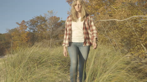handheld pan shot from behind dune grass to reveal a woman walking through the sand to the beach at sunset.  shot in 4k