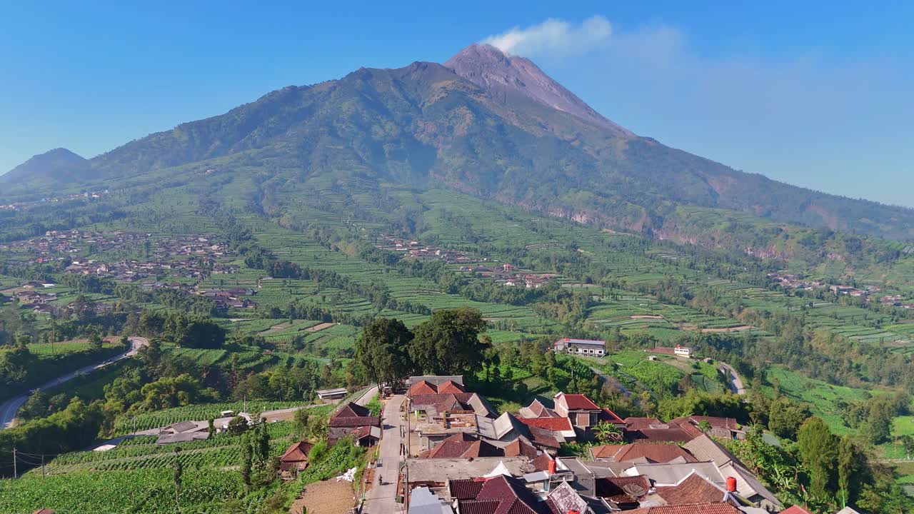 Vista aerea di un pittoresco villaggio rurale con un vulcano fumante circondato da una lussureggiante piantagione verde sullo sfondo sotto un cielo blu. Paesaggio rurale indonesiano con il vulcano Merapi.