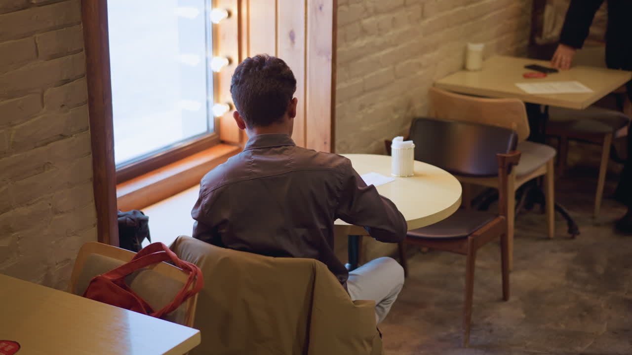 Man in formal shirt sitting with back to camera at small round table by window in cozy caf , writing on clipboard, coat draped on chair, soft lighting and warm brick wall create focused calm atmosphere