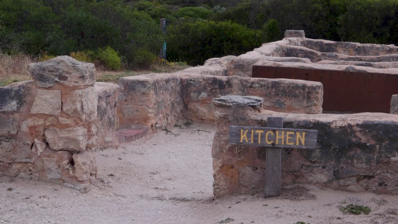 Un cartello logoro indica la zona cucina di un'antica struttura in pietra circondata da una vegetazione lussureggiante. Ideale per contenuti storici o educativi