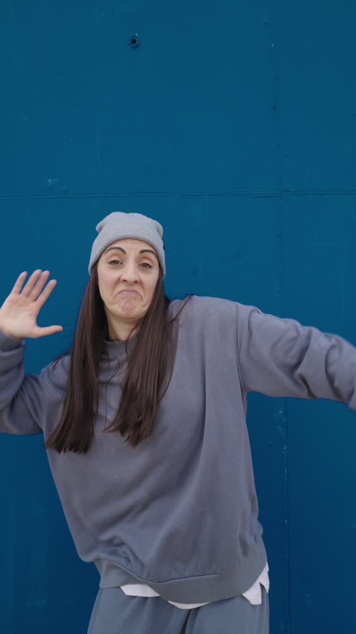 Woman in gray sweatshirt and beanie posing against blue background