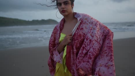 una mujer de cabello oscuro en un cardigan rojo y un vestido dorado de pie en la playa mirando fijamente a la cámara