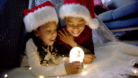 happy african american brother and sister in christmas hats playing with snow globe, slow motion